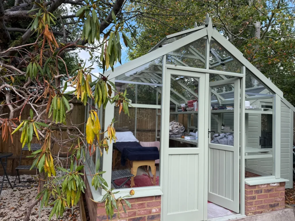 The sage-green glasshouse treatment room on a brick base in the garden, its door open to show the treatment bed