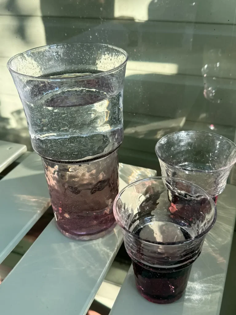 Hand-blown lilac glass tumblers of water on a slatted green shelf in the sunshine